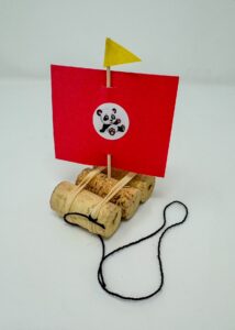 A completed boat. The body of the boat is comprised of three cylindrical corks fastened together with rubber bands. The boat has a red paper sail with a round panda sticker in the center, a yellow triangular flag at the top of the mast, and a black string. 